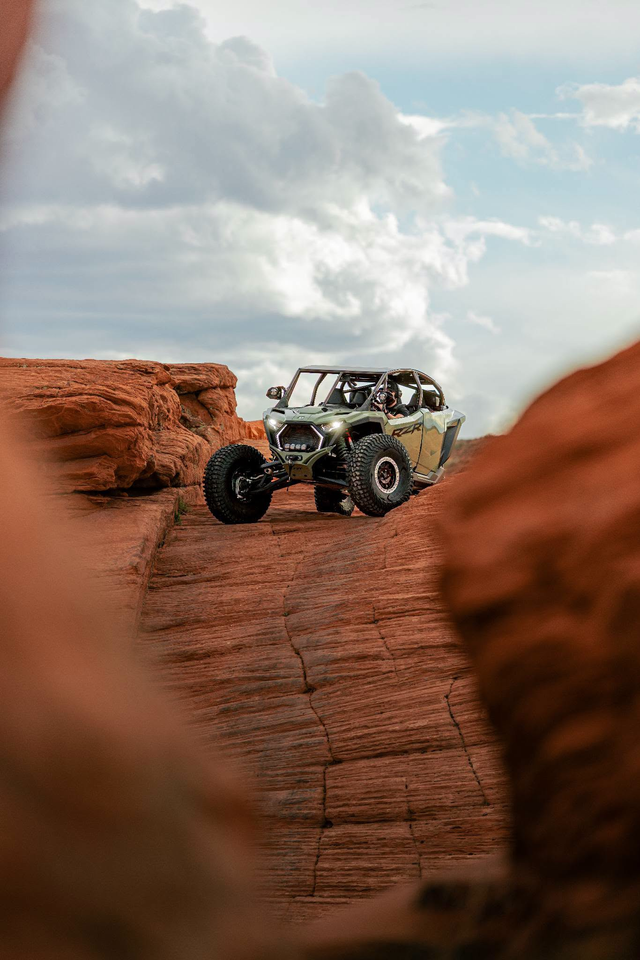 Polaris RZR with a helmeted driver navigating red sandstone terrain.